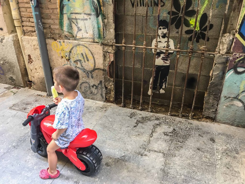das Foto zeigt einen Jungen mit einem Eis auf einem roten Plastikfahrzeug vor einem Stencil von Tona, das einen Jungen hinter einem Türgitter zeigt
