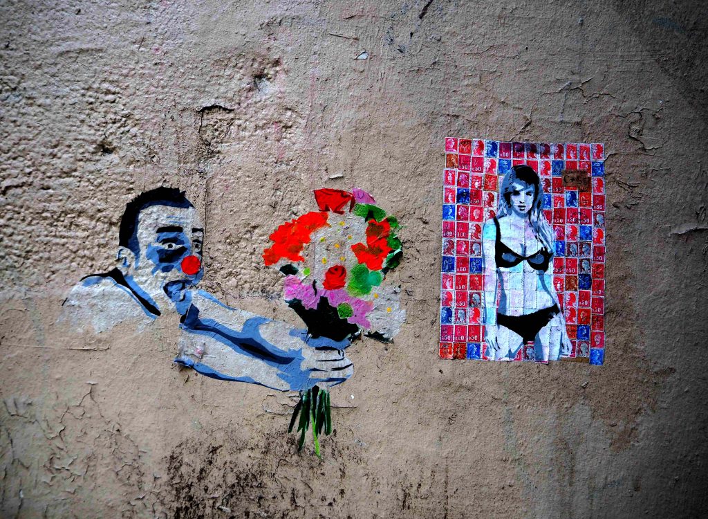 ein Clown streckt einer nur mit Bikini bekleidet jungen Frau einen Blumenstrauß entgegen